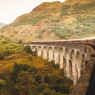 Looking from a window to the front of the train, which pulls carriages over the iconic Glenfinnan Viaduct towards a hillside.