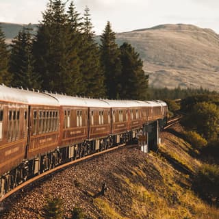 External view of the front ten carriages as the train heads towards hills, flanked by grassy escarpment and fir tree forest.