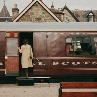 Viewed from behind, a woman in a cream coat uses a portable step to board the Royal Scotsman at Boat of Garten station.