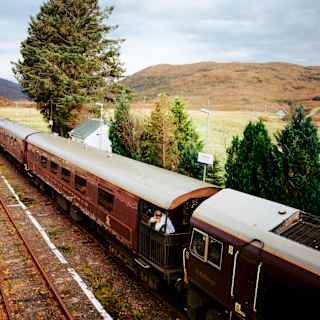 A man looks out from an observation deck as The Royal Scotsman waits at a rural station in the Highlands, seen from above.