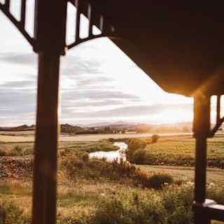 A silver river snakes through fields as the sun glows on the horizon, seen from the Observation Car deck, framed by railings.