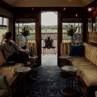 A woman relaxes with a drink on a banquette of warm gold upholstery as she gazes through the door of the observation deck.
