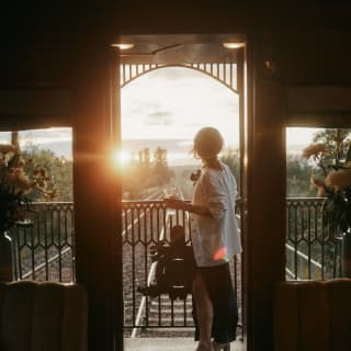 A woman in a white jacket watches the receding view with a sundown drink on the observation deck, seen from inside the car.
