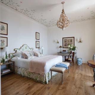 A double bed with fresh, white linen sits in the centre of a large airy suite with light walls and antique wooden furniture.