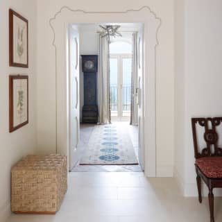Hallway with white walls and floor tiles and a door opening into a room with a grandfather clock and a sea-view window.
