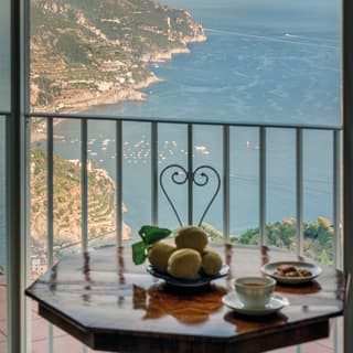 A polished antique table with fruit and coffee sits by the window with views on to a terrace and stunning Amalfi Coast.