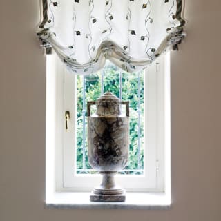 Detail of a window in a corridor of the villa, with a pretty, translucent blind and a decorative marble urn on the sill.