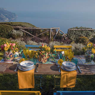 A rustic table is set for lunch in the sun overlooking the coastline, with blue and white bowls, yellow napkins and flowers.