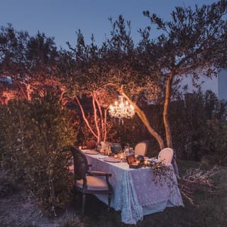 At dusk, a glittering chandelier hanging from the branches of an olive tree illuminates a table set for a celebration.