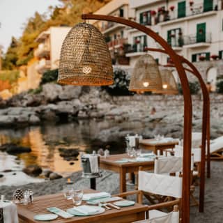 A row of teak dining tables extends along a private beach. A tulip-shaped lamp stand and champagne ice bucket serves each