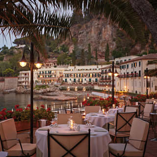 Tables set for dinner with crisp white tablecloths fill a terrace overlooking the bay. Candles flicker as dusk lowers