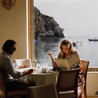 A couple study the breakfast menu as they sip coffee in the morning light. Behind them the Bay of Mazzarò looks like a painting