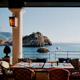 Elegant seaside restaurant terrace with laid tables and wicker chairs, overlooking calm blue water, rocky islands, and distant mountains under a clear sky. Decorative lanterns and potted plants frame the view.
