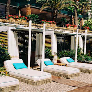 Three loungers with turquoise cushions recline in the sun outside cool Lido Villeggiatura cabanas with open drapes.