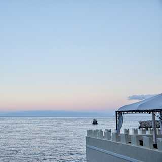 The soft pink of a sunset sky melt seamlessly into a silvery sea. In the foreground crenellations top the terrace wall