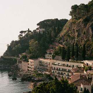 A coastal village with buildings nestled along a hillside, surrounded by trees and greenery, overlooking calm, rocky waters under a hazy sky.