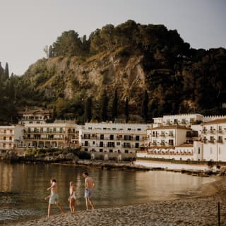 Three people play at the edge of the sand at Mazzarò beach as the hotel behind casts its reflections on the still waters.