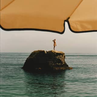 Surrounded by aquamarine waters, a man stands on a sea rock, saluting the camera, seen from beneath a yellow parasol canopy.