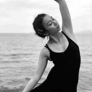 A woman in black yoga-wear and delicate necklace holds a leaning tree pose in a monochrome image with an ocean backdrop.