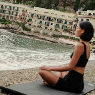 Angled soft-focus shot of a woman in black yoga wear, sitting in meditation pose on a mat on the beach in front of the hotel.