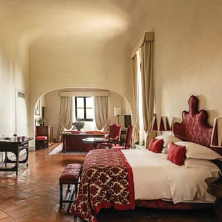 A carved stone fireplace faces a king bed, topped with red silk cushions and throw. An archway leads to a sitting room
