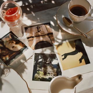 A marble table in dappled shade is strewn with polaroids, a porcelain cup and saucer of espresso, a plate and silver milk jug