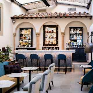 A stylish indoor bar with a tiled roof, two liquor shelves behind the counter, high barstools, and cushioned chairs around marble tables. The space is bright, with a glass ceiling letting in natural light.