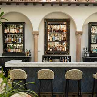 A stylish indoor bar with a marble countertop, four patterned barstools, and shelves stocked with bottles of spirits. Decorative arches and a plush sofa are visible in the foreground.