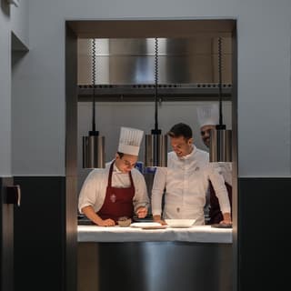Two chefs in chef whites and a red apron finishing a dish in a restaurant kitchen 