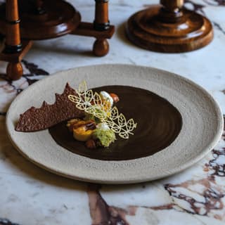 A plate of rich chocolate elements with green textured sponge cake, delicate tuiles and cocoa crisp, rests on a marble table.
