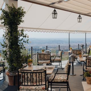 Dining tables and chairs rest in the shade of a wide canopy and enjoy panoramic views at the San Michele Grill by the Pool.
