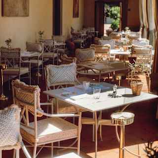 Sunlit outdoor café with empty tables and wicker chairs, set along a covered patio with potted plants and light streaming in from the side. The ambience is cosy and inviting, suggesting a peaceful place to relax.