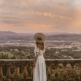 Lady in a white dress and sun hat standing on a rustic terrace gazing across the Florence skyline at sundown.