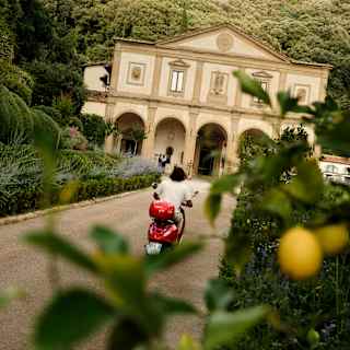 A man on a red vespa drives along the road towards Villa San Michele, in an angled shot taken from behind a lemon tree.