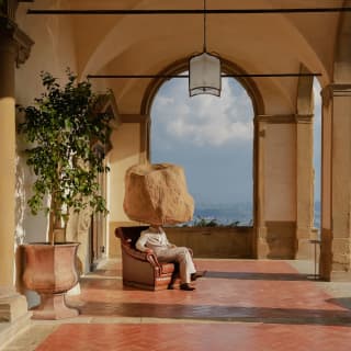 In an art installation a man sits on a leather chair in a stone columned loggia. In place of his head is a sandstone boulder