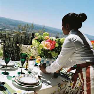 A woman in a white shirt and striped apron lays a table outdoors with floral arrangements, plates, and green glasses, overlooking a scenic landscape under a clear blue sky.