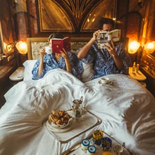 A couple in blue gowns read in bed with two breakfast trays nestled between them, with coffee, pastries, juice and yoghurt.