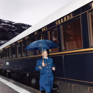 A steward in blue coat with gold buttons and cuff stripes smiles as he dashes through the rain below a matching blue umbrella