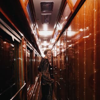 A woman in an evening gown smiles back over her shoulder as she walks along a narrow wood panelled train corridor