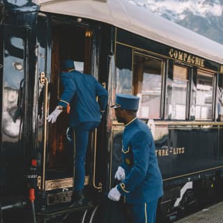 Two stewards in blue suits with gold trim, flat topped hats and white gloves board the train with the Alpine peaks behind