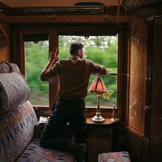 With a knee resting on a tapestry-like upholstered seat, a man stands at an open window, taking in the view from the train