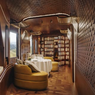 A lounge view glances a window-side daybed with book and binoculars into a calm space with yellow chairs, table and library.
