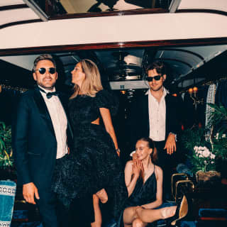 There is a sense of carousing in the air as two men in black tie and sun glasses laugh with two women in black cocktail dresses