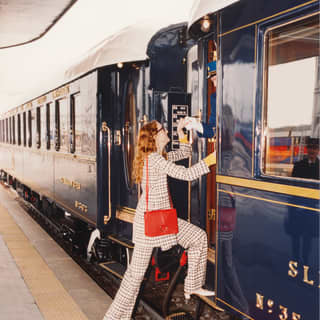 Lady in an elegant checkered trouser suit stepping from a red carpet on to a vibrant blue vintage train carriage