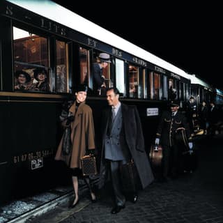 A couple dressed elegantly in coats and hat walk by the side of the carriage. Behind them gold-trimmed stewards carry bags