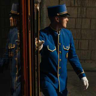 A man in a blue Pullman Porter uniform with gold braiding and a kepi hat stands in the doorway of the stationary train.