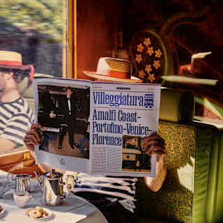 A man in a striped gondolier shirt and hat holds up a Villeggiatura paper, seated by train window which reflects his profile.