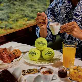 A person in a blue patterned shirt drinks espresso at a breakfast table with pastries, orange juice, fruit, and a plate of tennis balls labelled Caviar. A window shows greenery outside.