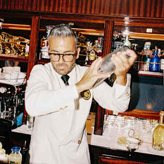 A barman in a white jacket and black tie is shaking a cocktail shaker behind a wooden bar, with shelves of bottles and glasses in the background.
