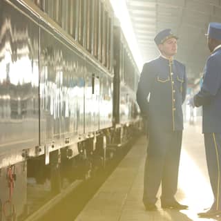 2 stewards in blue and gold uniforms share information beside the train on a platform bathed in a shard of early morning light